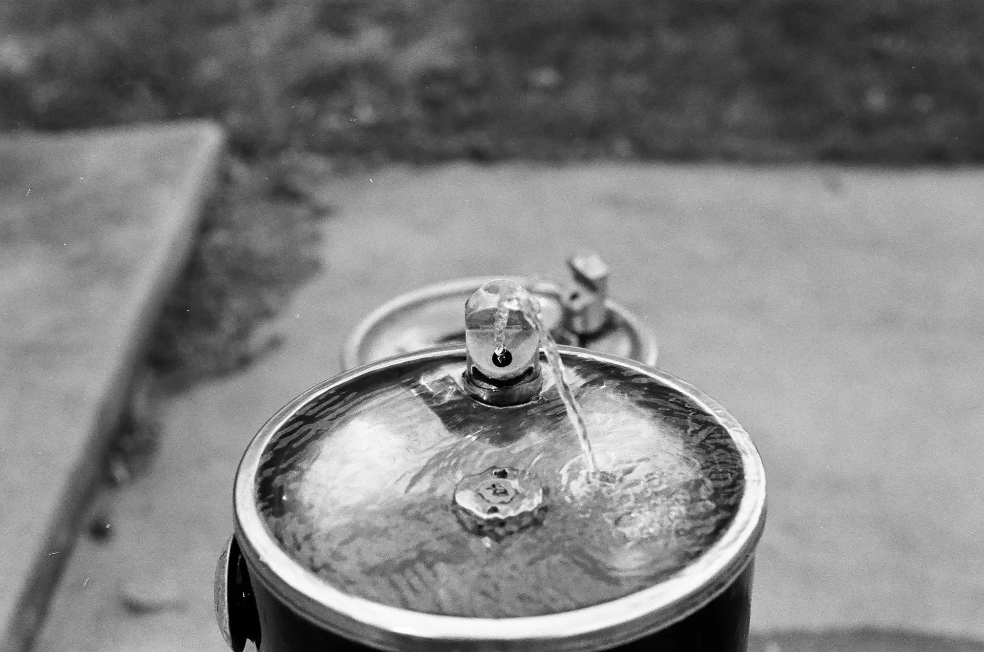 Black-and-white photograph of a circular water fountain. its bowl is full up with water. With no one operating it, one can assume that it's set to always be on. The arc of its output bends to the right in the wind.