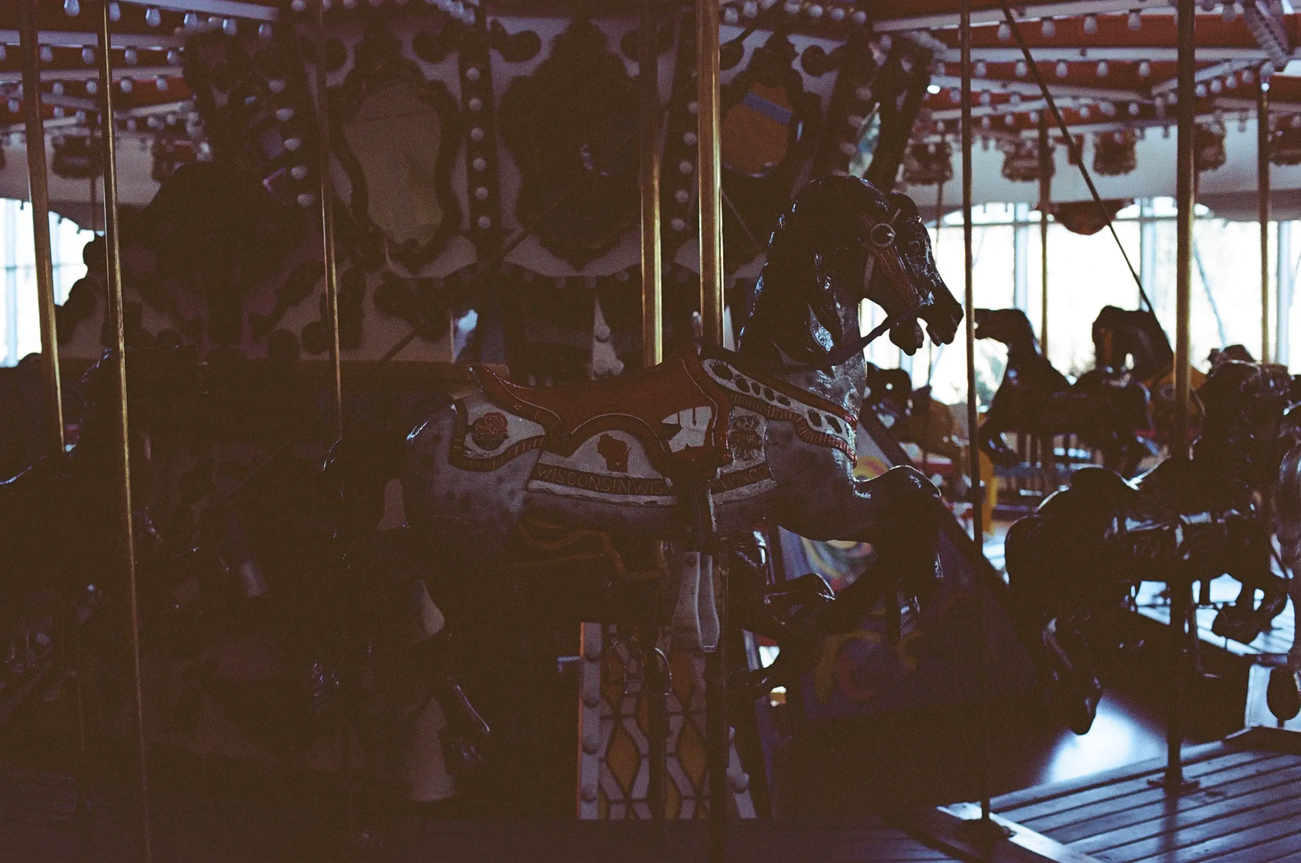 A dark photograph of a carousel horse. No power runs through the machine; it's lined with lightbulbs but none are on. The horse is adorned in orange and white, its saddle reading "WISCONSIN". Some spots mark its gray fur.