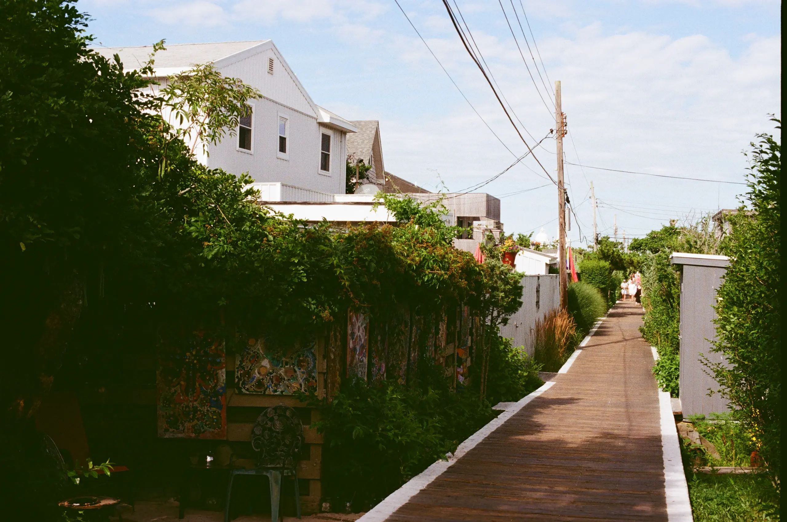 Photograph of a wood boardwalk lined with white stretching into the distance. On all sides are houses -- in particular, a large all-white house is clearly visible above fences. In the dark shade of trees is a set of mosaic artworks.