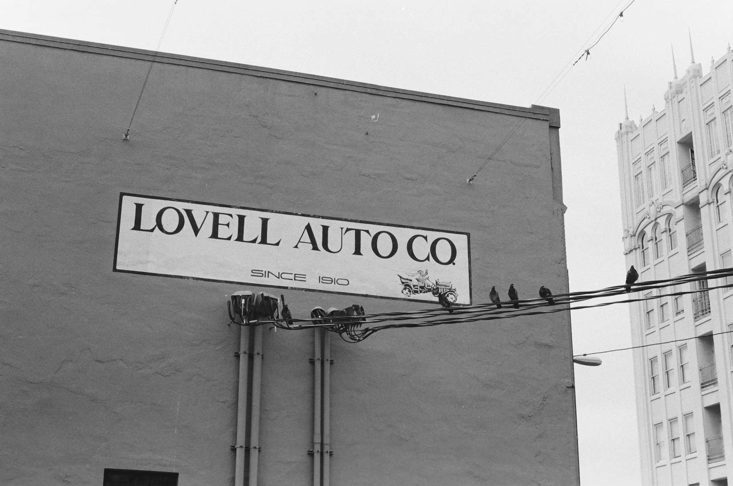 Black-and-white photograph of a gray, largely featureless building. It's adorned with painted-on signage, reading "LOVELL AUTO CO. SINCE 1910". Reaching away from this label is a bundle of power cables, atop which sits a group of pidgeons.