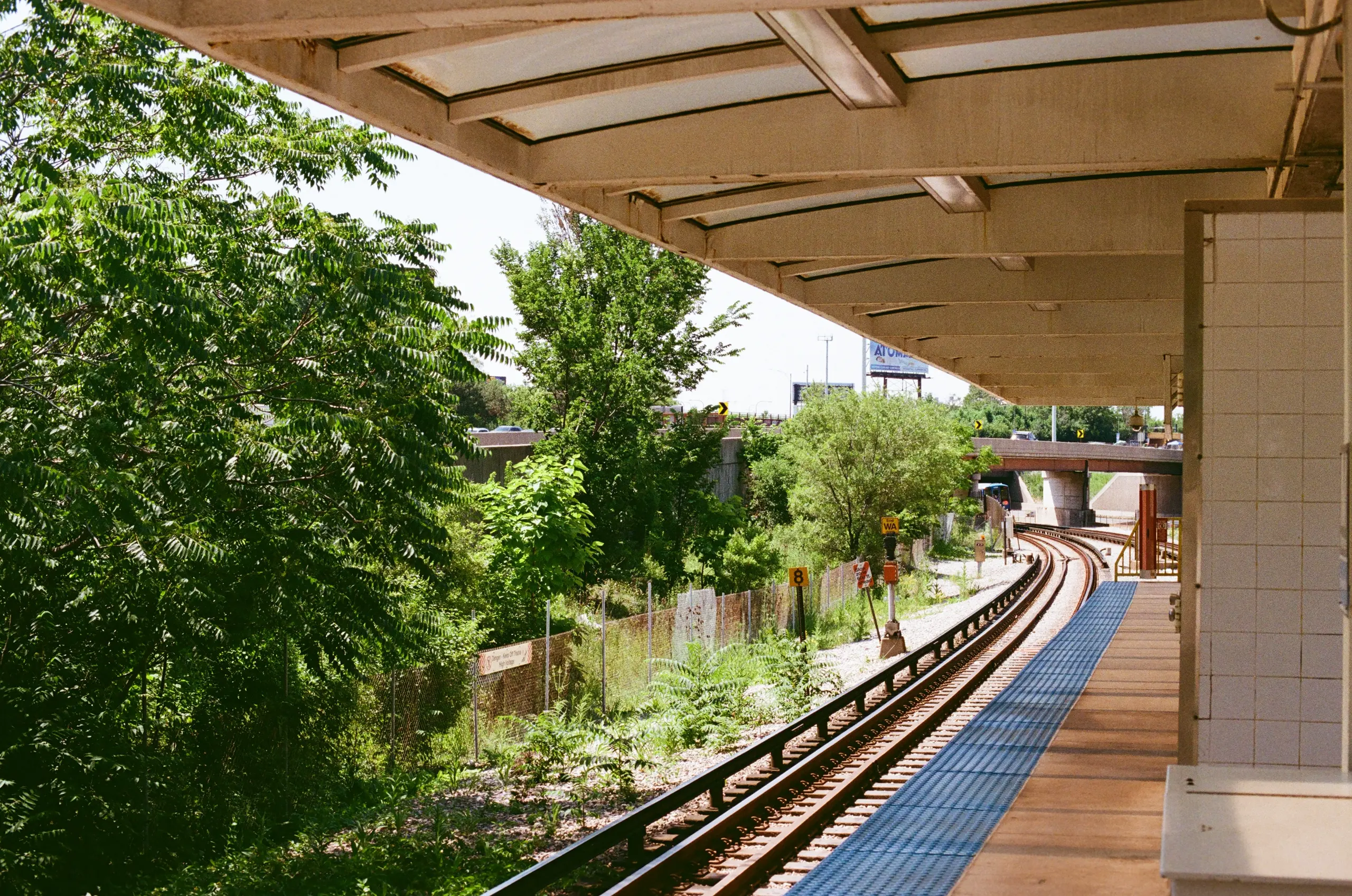 Photograph of a train track winding away from a boarding platform and underneath a bridge. Across from the roofed platform are trees, blocked off by an orange, plastic-mesh fence. In the distance is raised a billboard.