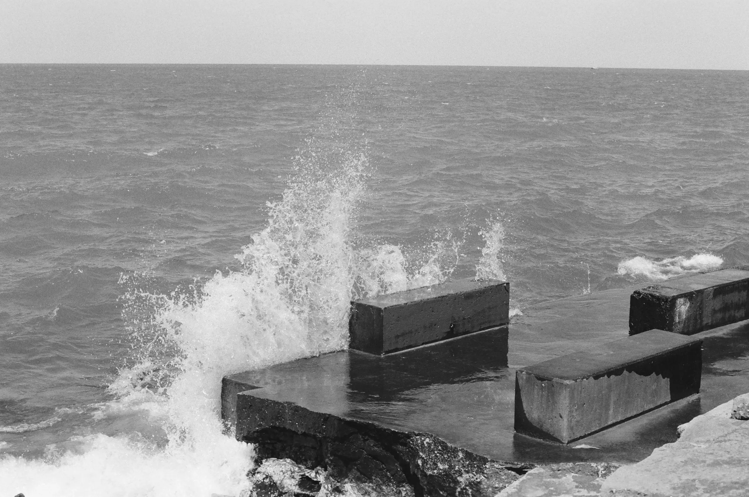 Black-and-white photograph of water crashing against a concrete structure. The concrete is mostly smooth, marked only by purpose-built rectangular protrusions and erosion-related wear-and-tear. The bright white of the splash contrasts form the grays of the concrete and lake.