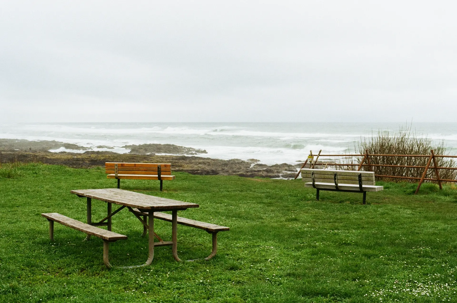Photograph of two benches and a picnic table on bright green grass. Past the benches is a ledge overlooking a rocky beach. The ocean is a light gray-blue, with white capped waves rolling towards the shore. Some crash against the rocks.