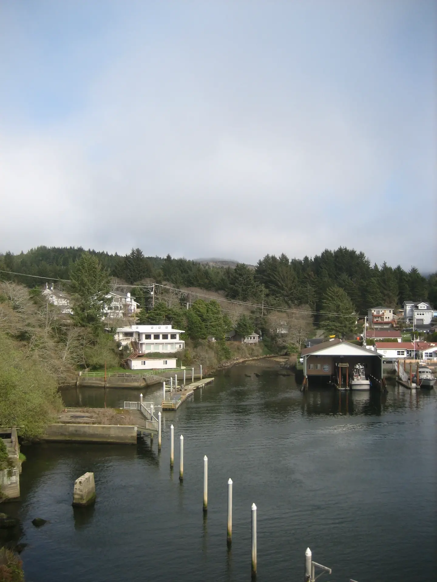 Photograph of a small-town waterfront on a not-quite-cloudy day. A small hill runs in a curve along the water, lined densely with trees and sparsely with buildings, mostly white. Near to the edge is various seafaring paraphernalia -- docks, white pillars, and a few boats.