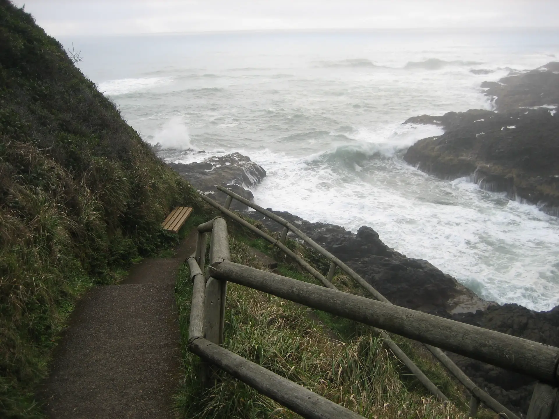 A photograph staken from the top of a steep trail leading down to rough waters crashing and sliding from painful, uneven rocks. The trail makes an upside-down "V", each edge lined with an old-but-sturdy log rail and a steep, wild-grass hill. At the "V"'s joint lives a new-looking bench, light brown and nested in plants.
